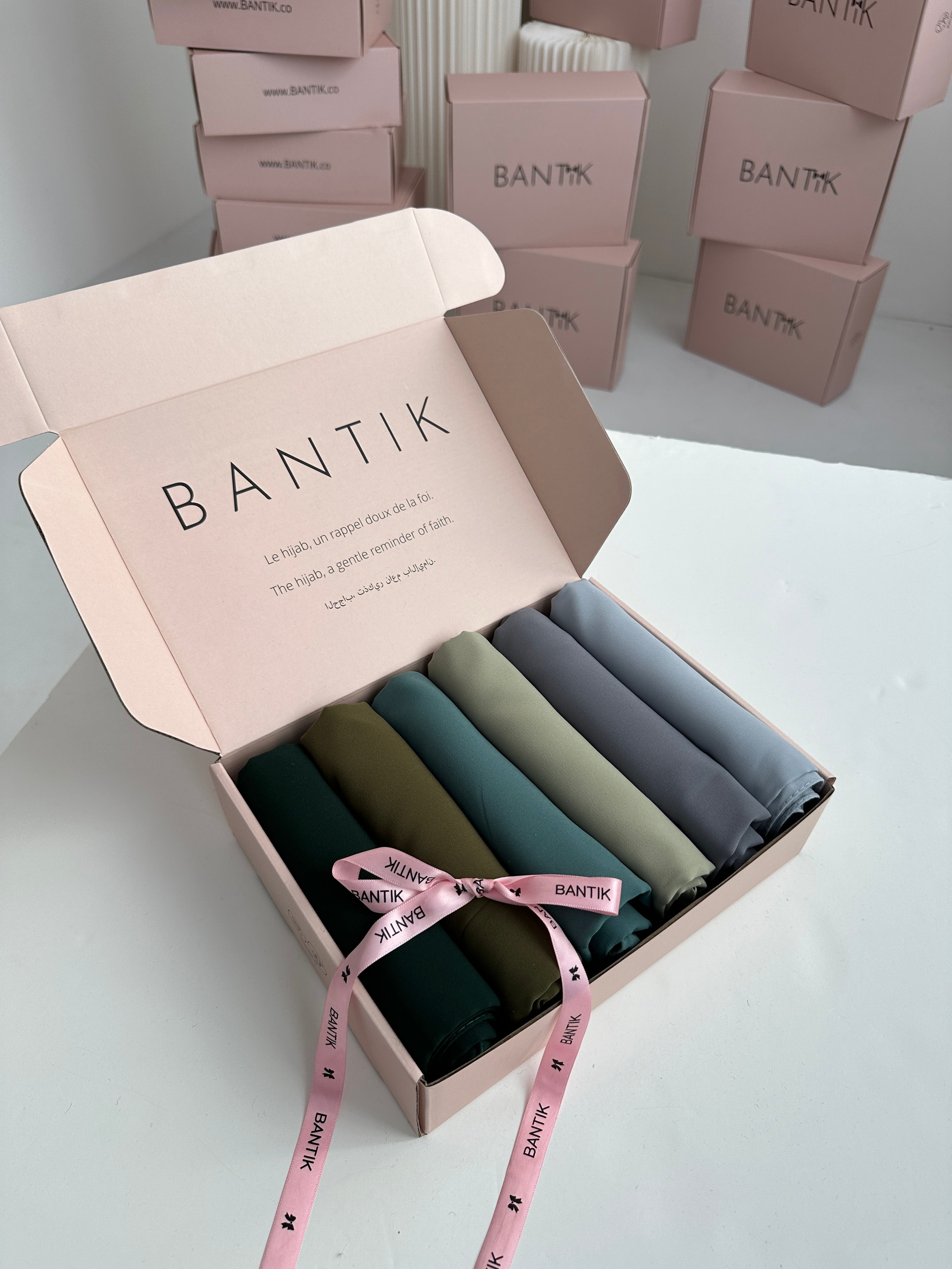 Coffret cadeau ouvert contenant 6 hijabs en Soie de Médine, roulés et alignés, dans une palette de couleurs dégradées allant du mauve foncé au lilas clair et beige rose. Le coffret est noué par un ruban rose marqué 'BANTIK'.