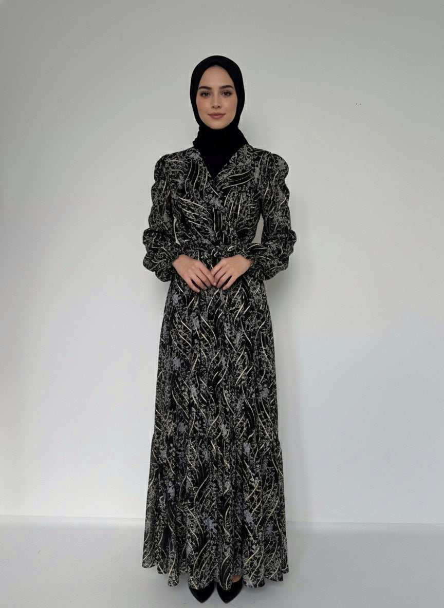 Robe longue en mousseline noire avec un imprimé doré et gris abstrait, portée sur un mannequin. La robe a un col en V, une ceinture à nouer à la taille et des manches longues bouffantes avec des poignets élastiques. Un hijab noir couvre la tête du mannequin.