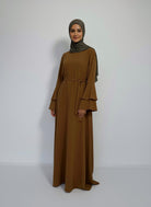 Robe longue marron ocre en tissu fluide, présentée sur un mannequin. La robe a un col rond, des manches longues avec des volants étagés au niveau des poignets, et une ceinture amovible à la taille.
